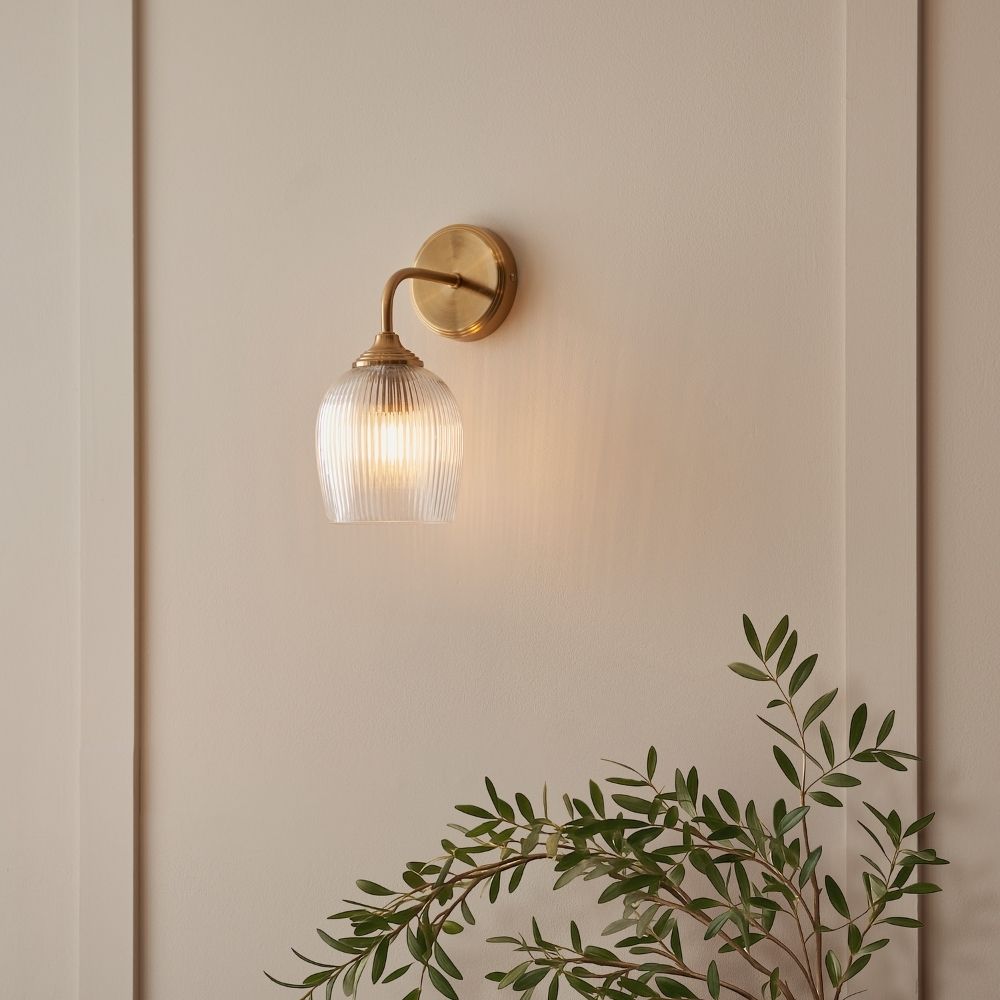 Aged brass wall light with ribbed glass shade, creating a warm ambient glow on a neutral interior wall.