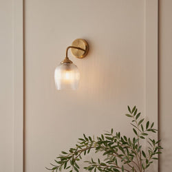 Aged brass wall light with ribbed glass shade, creating a warm ambient glow on a neutral interior wall.