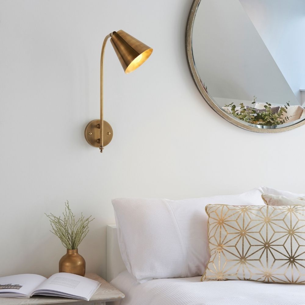 Antique brass adjustable wall reading light installed beside bed with decorative cushions and mirror in modern interior setting.