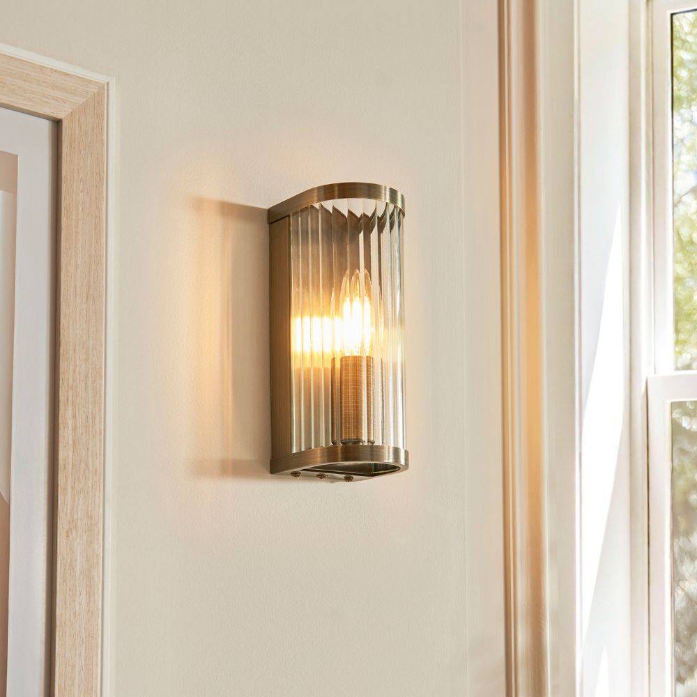 Antique brass wall light with a ribbed clear glass shade and exposed candle-style bulb for elegant ambient lighting.