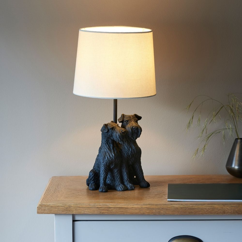 Black resin table lamp featuring two sitting dogs with textured detail and a neutral fabric lampshade, styled on a wooden bedside table.