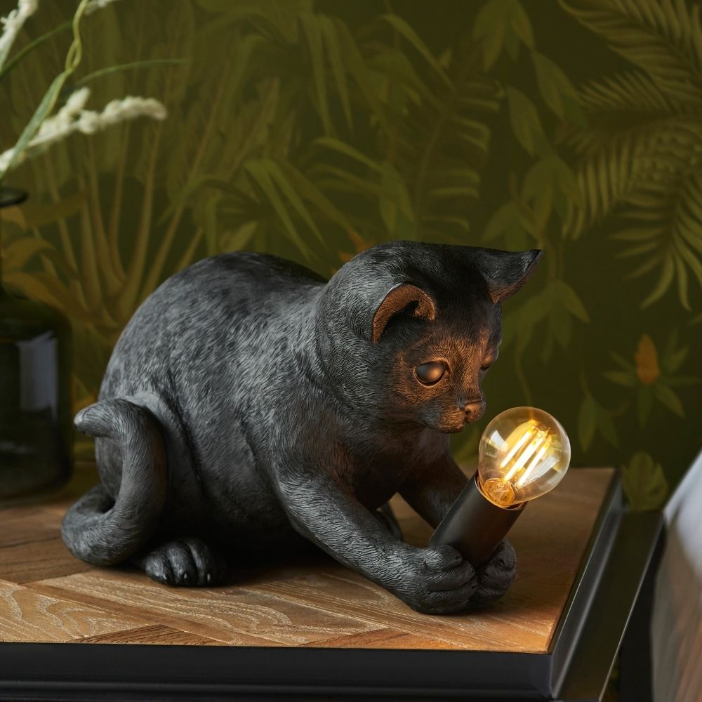 Black kitten resin table lamp holding exposed filament bulb, styled on wooden surface with warm ambient lighting and botanical background.