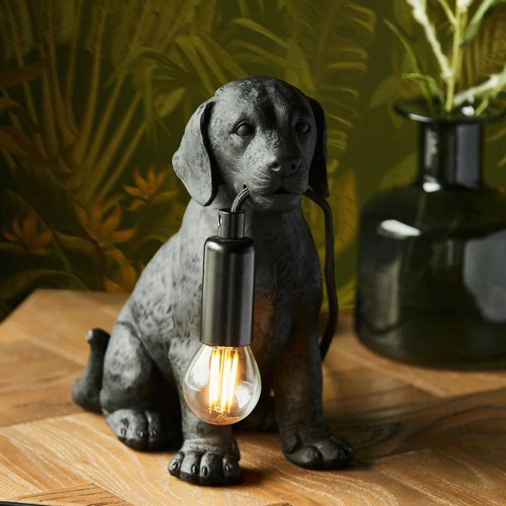 Black Labrador puppy resin table lamp holding exposed filament bulb, styled on a wooden surface with warm ambient lighting and botanical background.