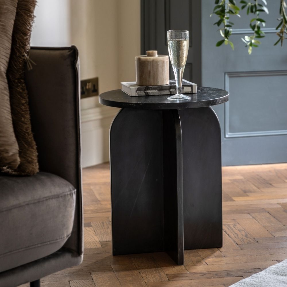 Black marble sculptural side table styled beside an armchair with books and a glass on top in a contemporary living room.