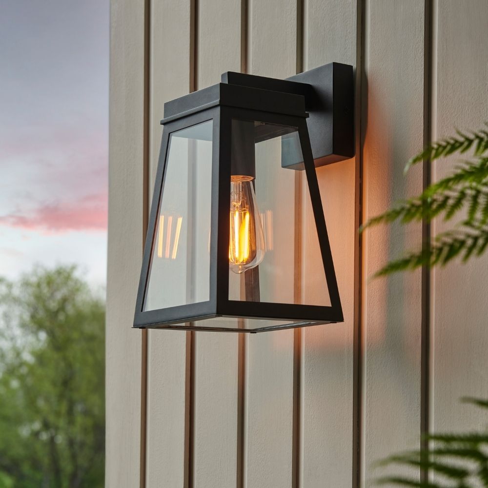 Black modern outdoor lantern wall light with clear glass panels mounted on exterior wall with warm glowing filament bulb.