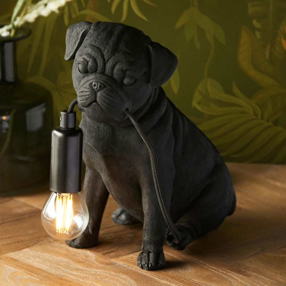 Black pug puppy resin table lamp holding exposed filament bulb in its mouth, styled on a wooden surface with warm ambient lighting and botanical background.