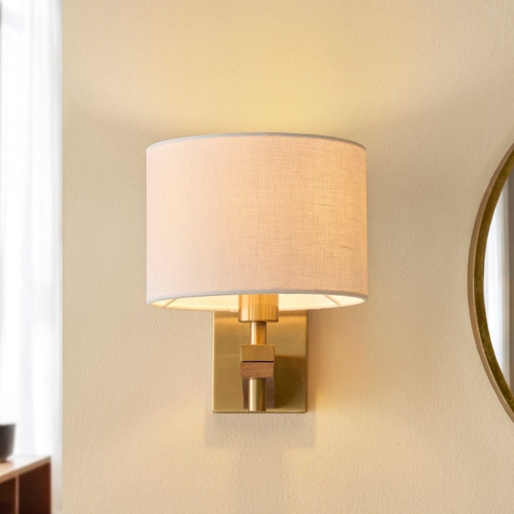 Brass and ash wood wall light with linen shade illuminating a contemporary interior beside a mirror and seating area.