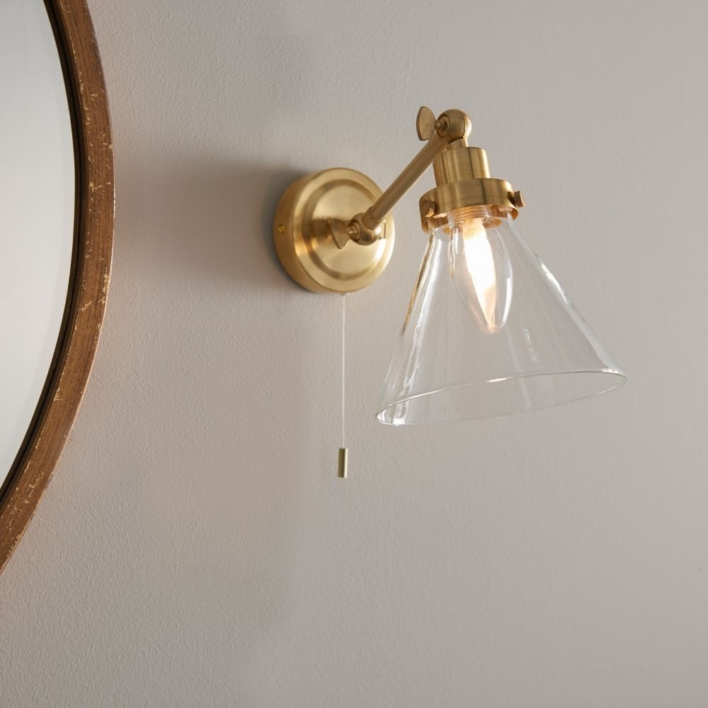 Brass wall light with adjustable arm and clear glass shade mounted beside a round mirror in a modern bathroom.