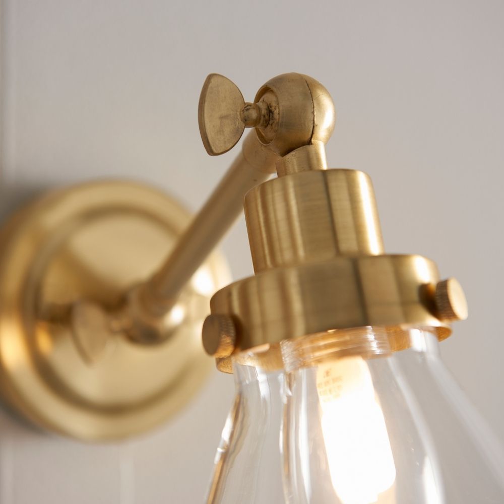 Close-up detail of brass wall light showing adjustable arm joint, fittings and illuminated bulb inside clear glass shade.