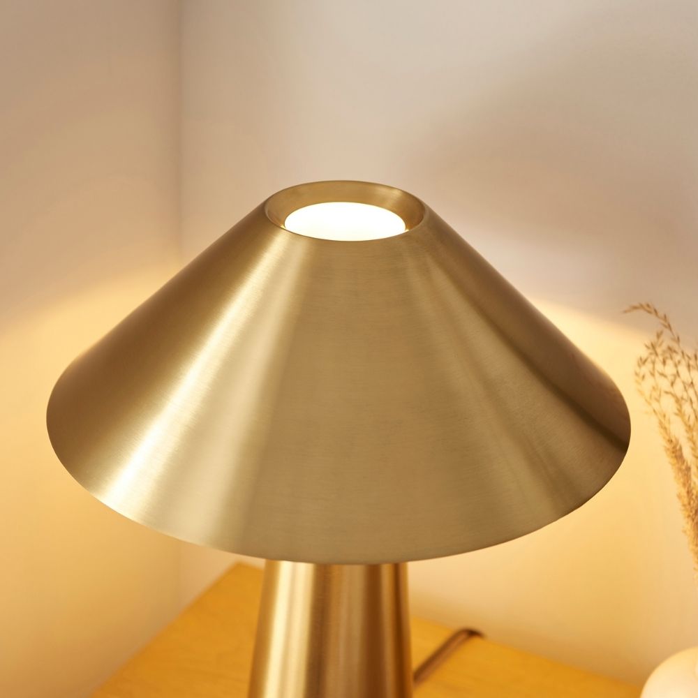 Modern brushed brass table lamp with a wide cone-shaped shade and sleek cylindrical base, emitting warm ambient light.
