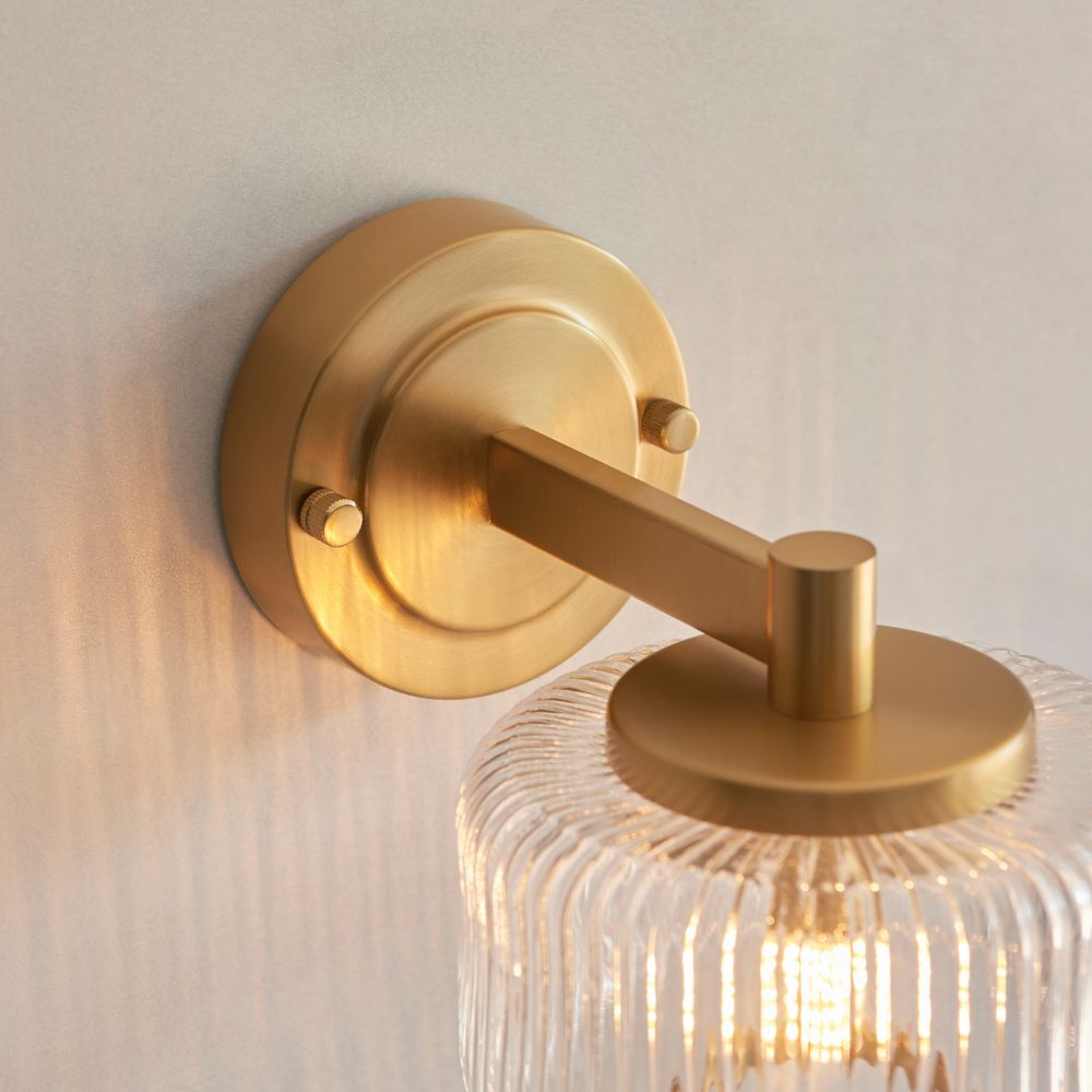 Brushed brass wall-mounted light with a clear ribbed glass shade and modern minimalist styling.