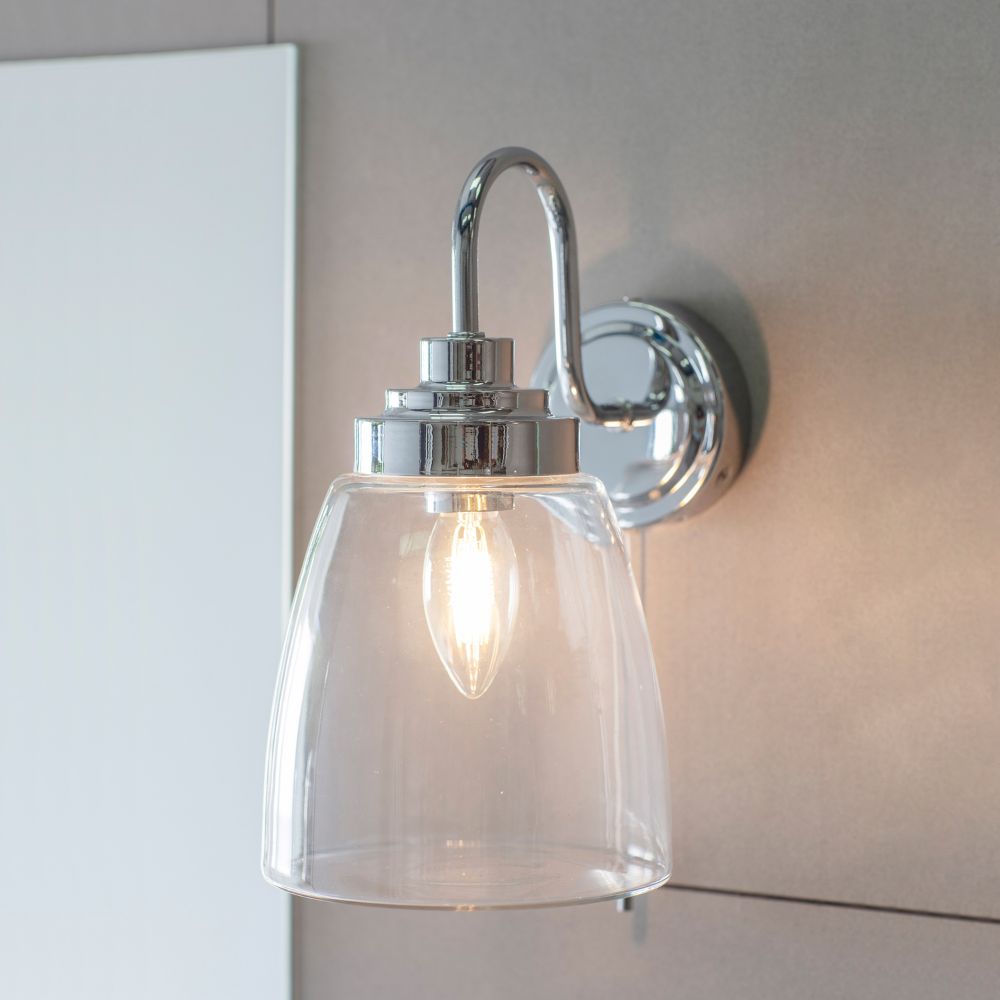 Modern chrome wall light with clear glass shade installed beside a bathroom mirror and illuminated with a warm LED bulb.