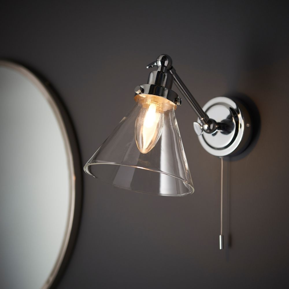 Chrome wall light with adjustable arm and clear glass shade, mounted beside a mirror in a modern bathroom setting.