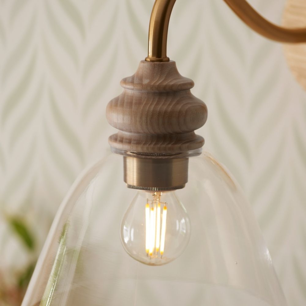 Clear glass shade wall light with turned ash wood detail and curved aged brass arm on a neutral background.