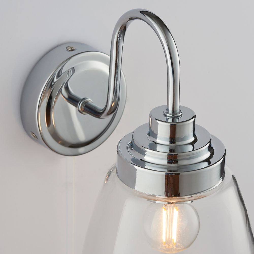 Detailed close-up of a polished chrome wall light fitting with illuminated bulb and clear glass lampshade.