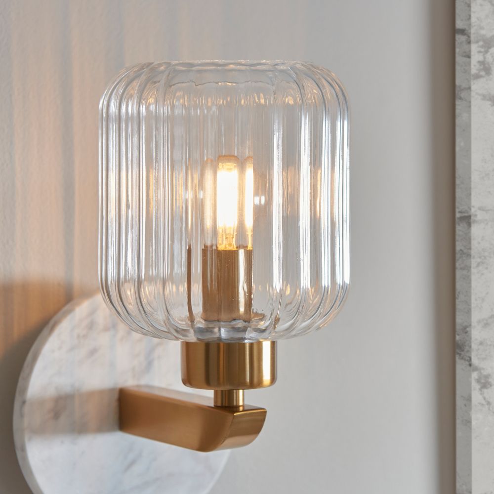 Close-up of a ribbed clear glass wall light with brushed brass finish and softly illuminated bulb for modern interiors.