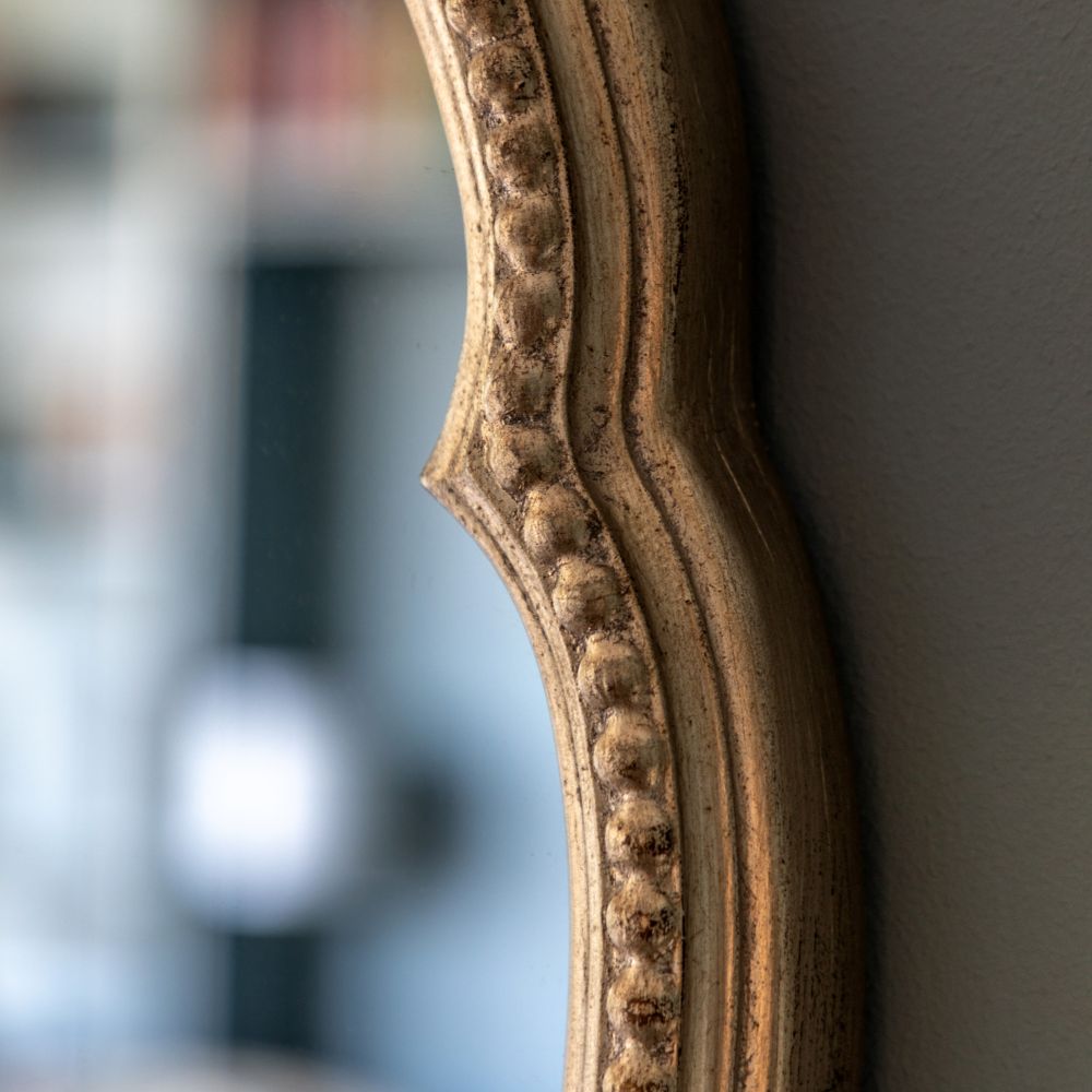 Angled close-up of an ornate round mirror frame with scalloped edging, antique gold finish and decorative beaded detailing.