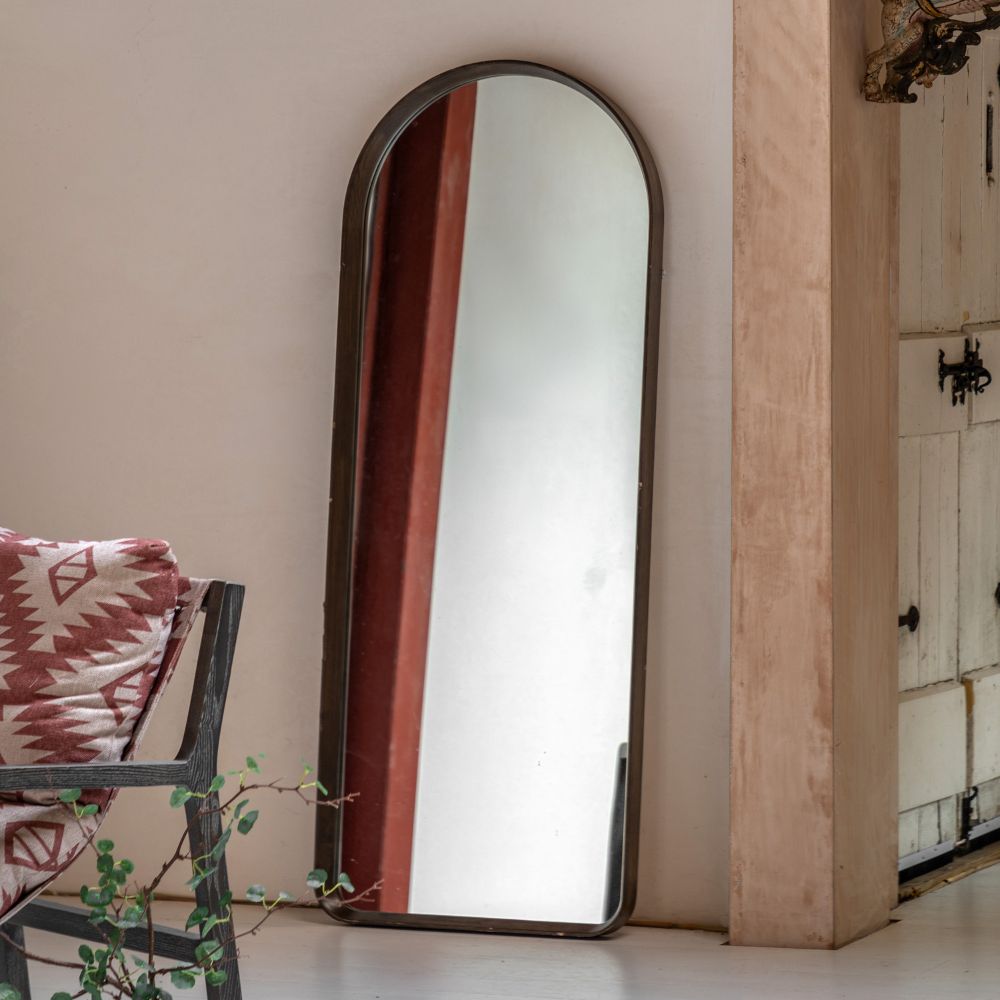Arched full length floor mirror with a dark stained wooden frame leaning against a wall in a rustic interior beside a patterned chair and wooden beams.