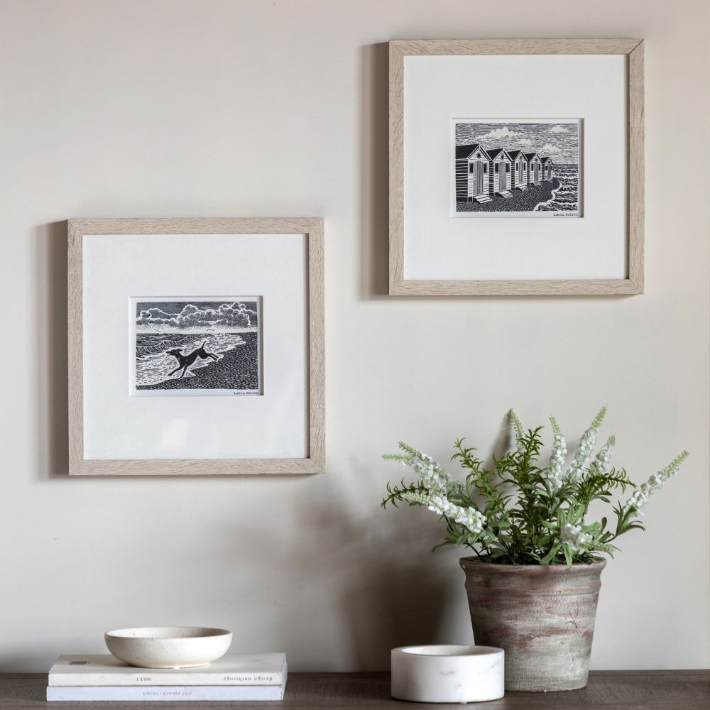 Set of two framed coastal linocut prints featuring beach huts and a seaside dog scene in monochrome illustration style.