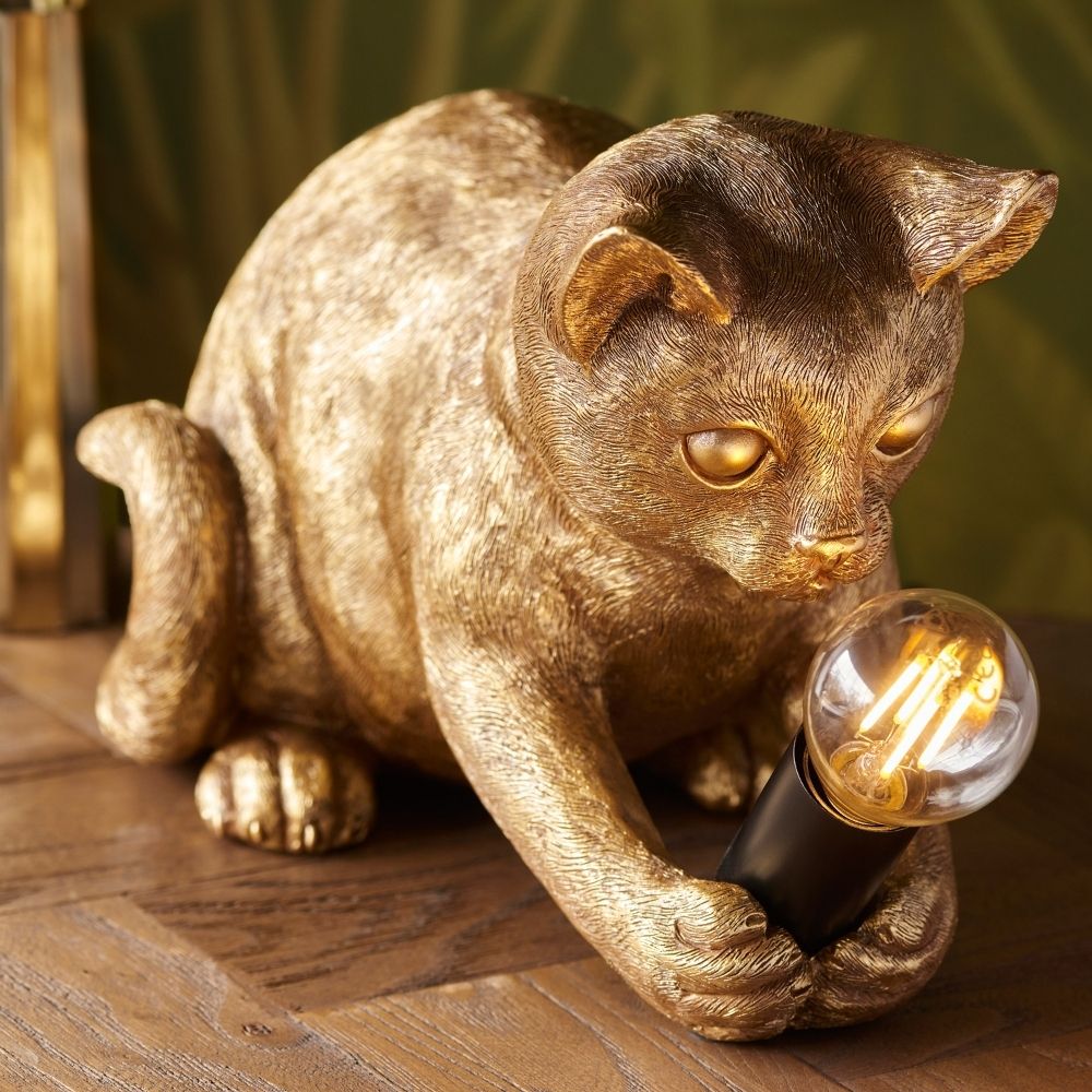 Gold cat resin table lamp holding exposed filament bulb, featuring decorative lighting design with textured finish on wooden surface.