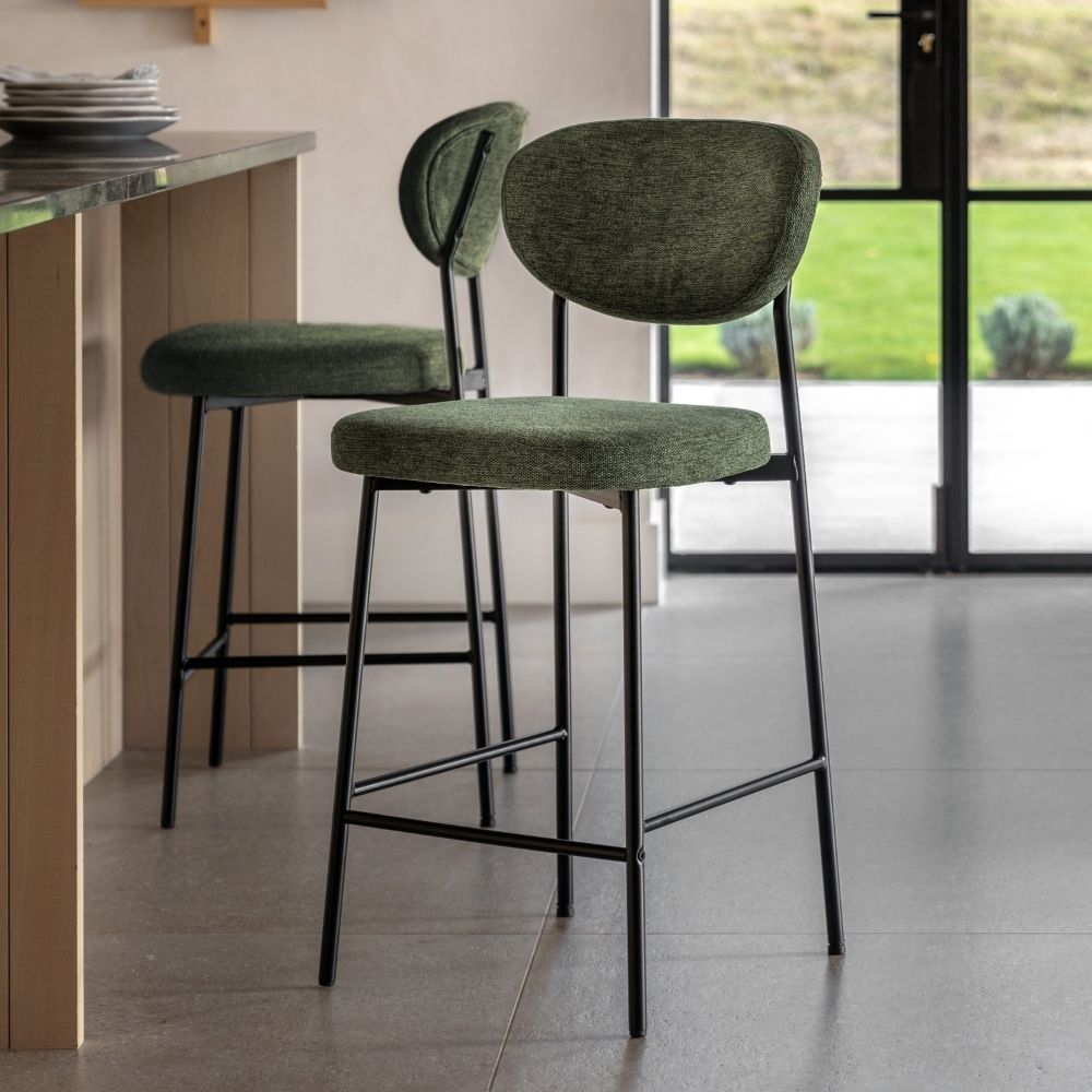 Two green fabric upholstered bar stools with black metal legs placed in a modern kitchen interior beside a breakfast bar and glass doors.