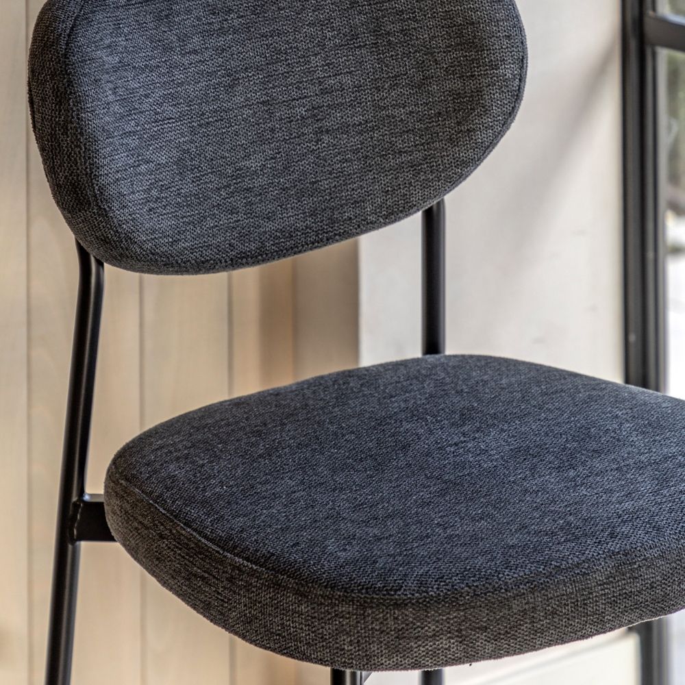 Close-up of a grey fabric bar stool showing the padded seat, curved backrest and black metal frame in an interior setting.