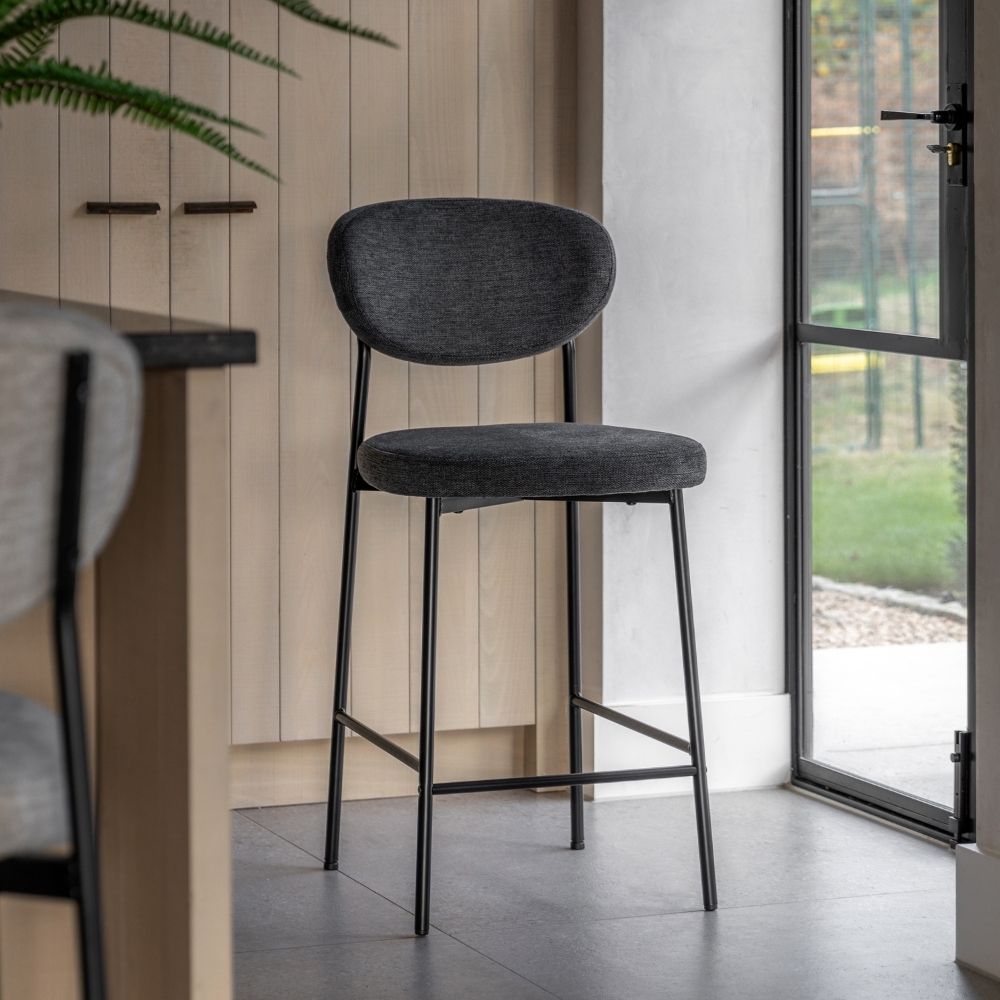 Grey fabric upholstered bar stool with black metal legs placed in a modern kitchen beside a glass door and light wood cabinetry.