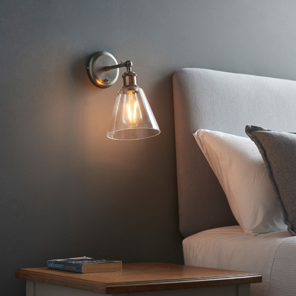 Industrial bedside wall light with a clear glass shade and exposed warm LED bulb mounted beside a modern upholstered bed.