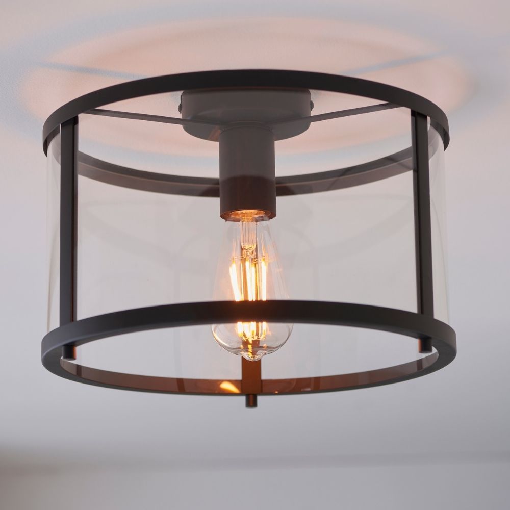 Industrial black semi flush ceiling light with cylindrical metal cage and clear glass shade surrounding an exposed filament bulb.