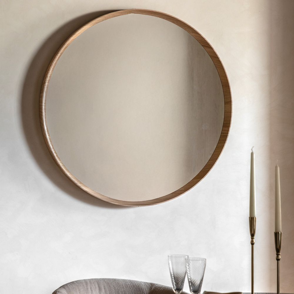 Large round wall mirror with a natural oak wooden frame styled above a modern dining table with candlesticks and glassware in a neutral interior.