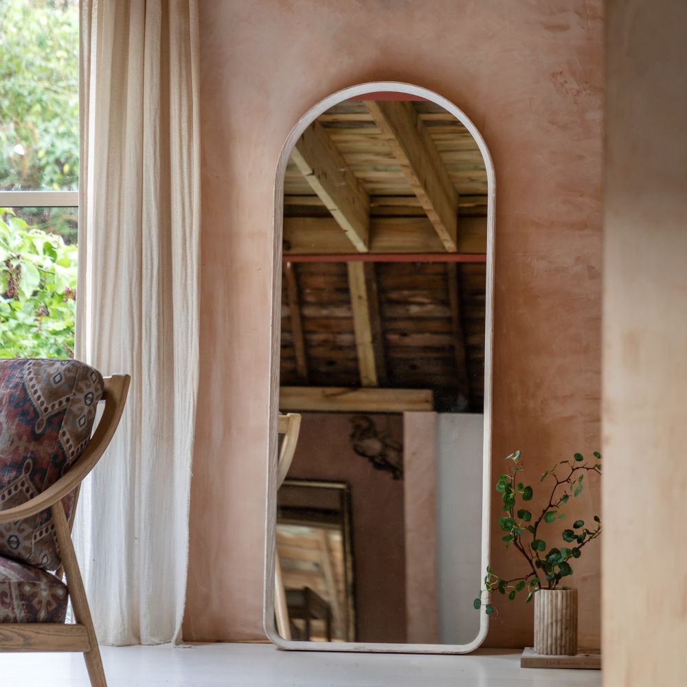 Light oak arched full length floor mirror leaning against a textured wall in a rustic interior with soft curtains and decorative greenery.