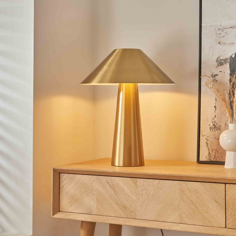 Minimalist brass table lamp with conical shade placed on a wooden sideboard, creating soft warm lighting in a contemporary interior.