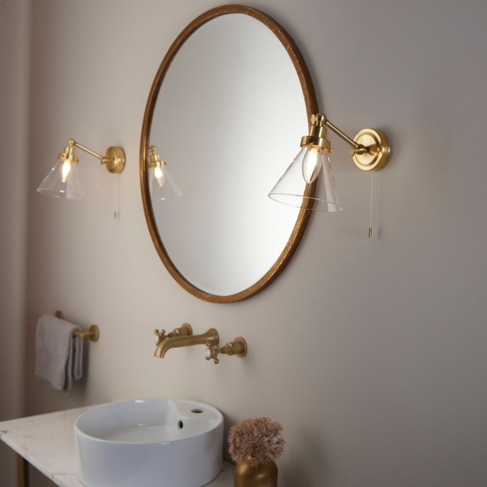 Pair of brass wall lights with clear glass shades installed either side of a round mirror above a bathroom sink in a contemporary setting.