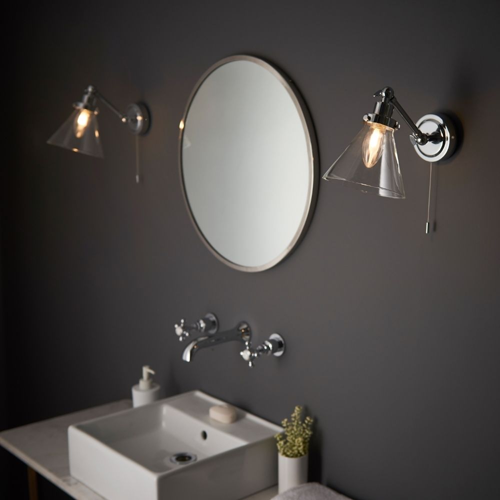 Pair of chrome wall lights with clear glass shades installed on a bathroom wall either side of a round mirror above a sink.