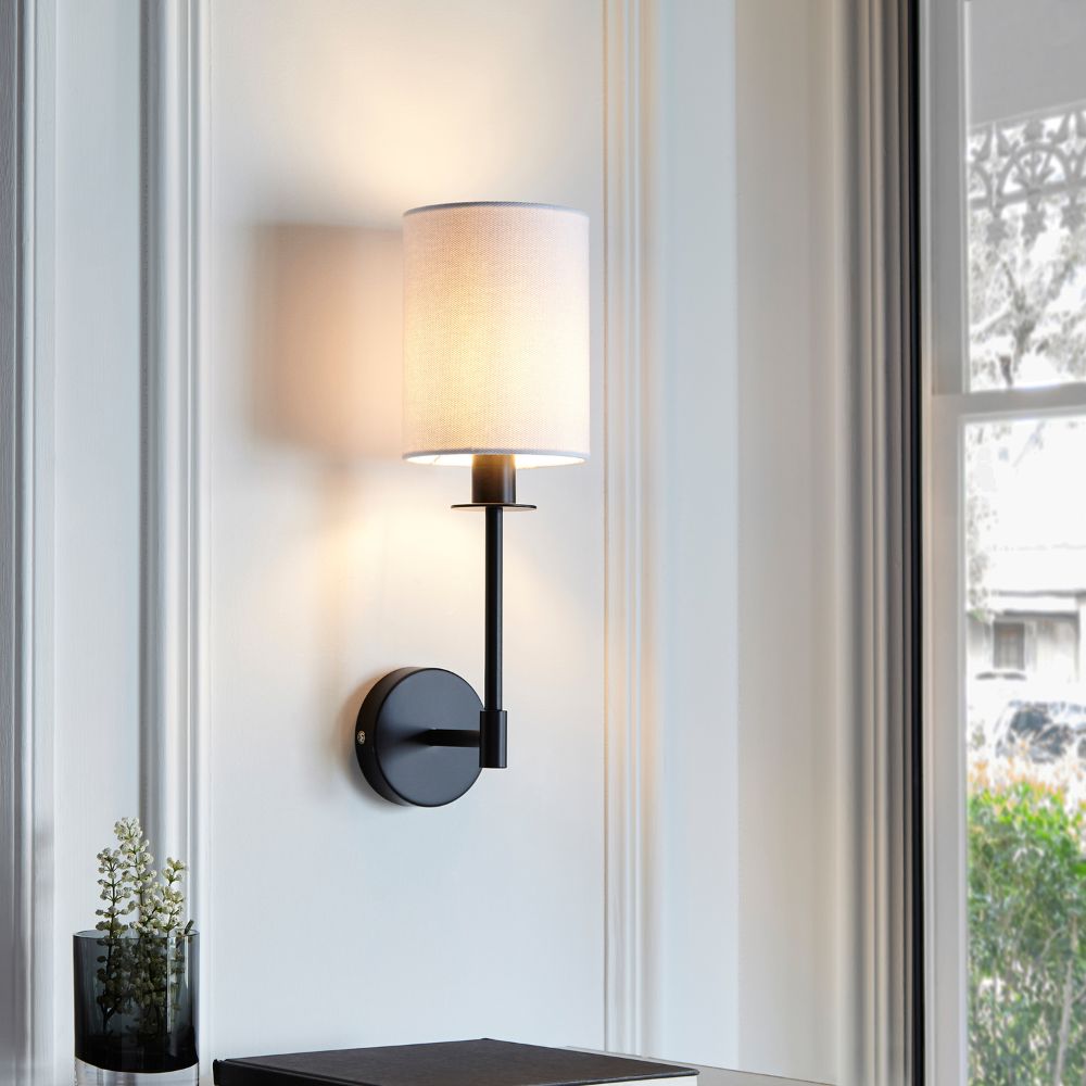 Modern bedroom wall sconce with a white cylindrical fabric shade and black metal frame mounted beside a window.