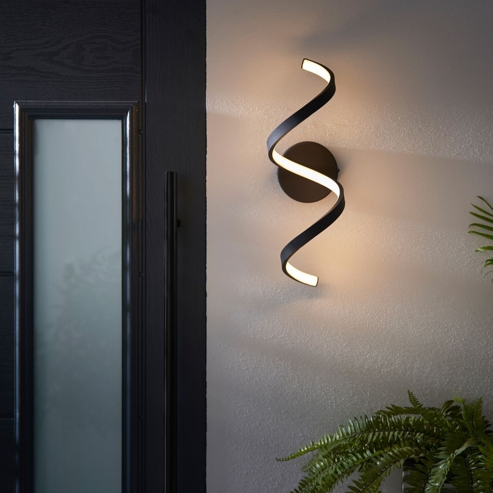 Black twisted LED wall light installed beside a door with soft ambient lighting and outdoor greenery nearby.