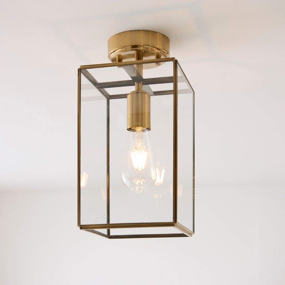 Modern ceiling light fixture with a brass frame and clear glass panels enclosing a visible filament bulb.