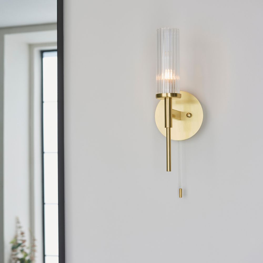 Modern brass wall light with a ribbed clear glass cylinder shade and elegant pull cord switch for ambient interior lighting.