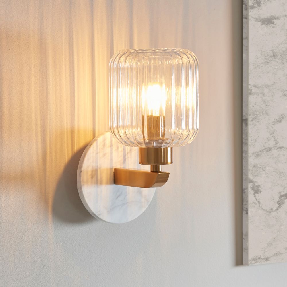 Modern brass wall light with a ribbed clear glass shade and white marble-effect backplate casting a warm ambient glow.