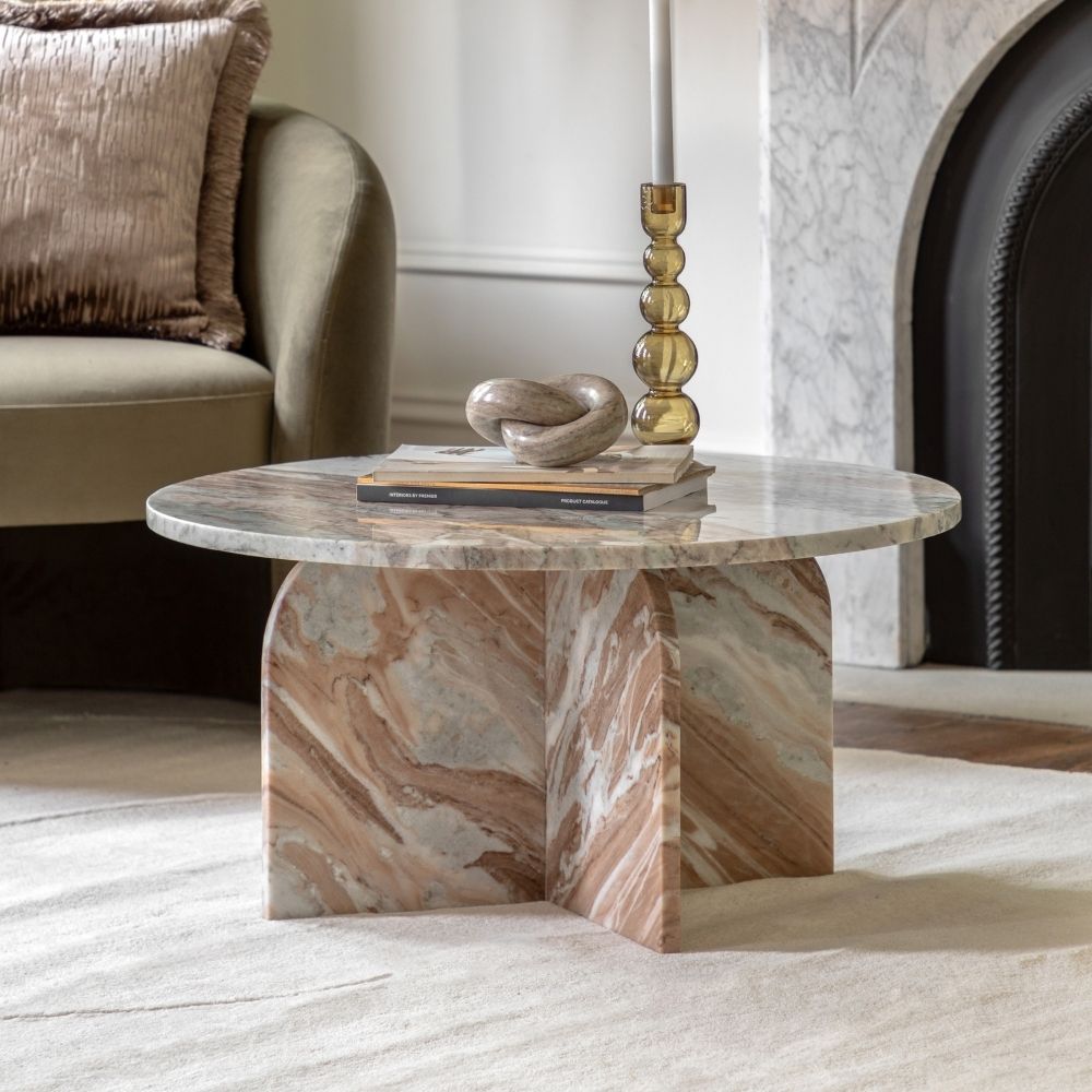 Round natural marble coffee table with sculptural base styled in a cosy living room beside an armchair and fireplace with decorative accessories.