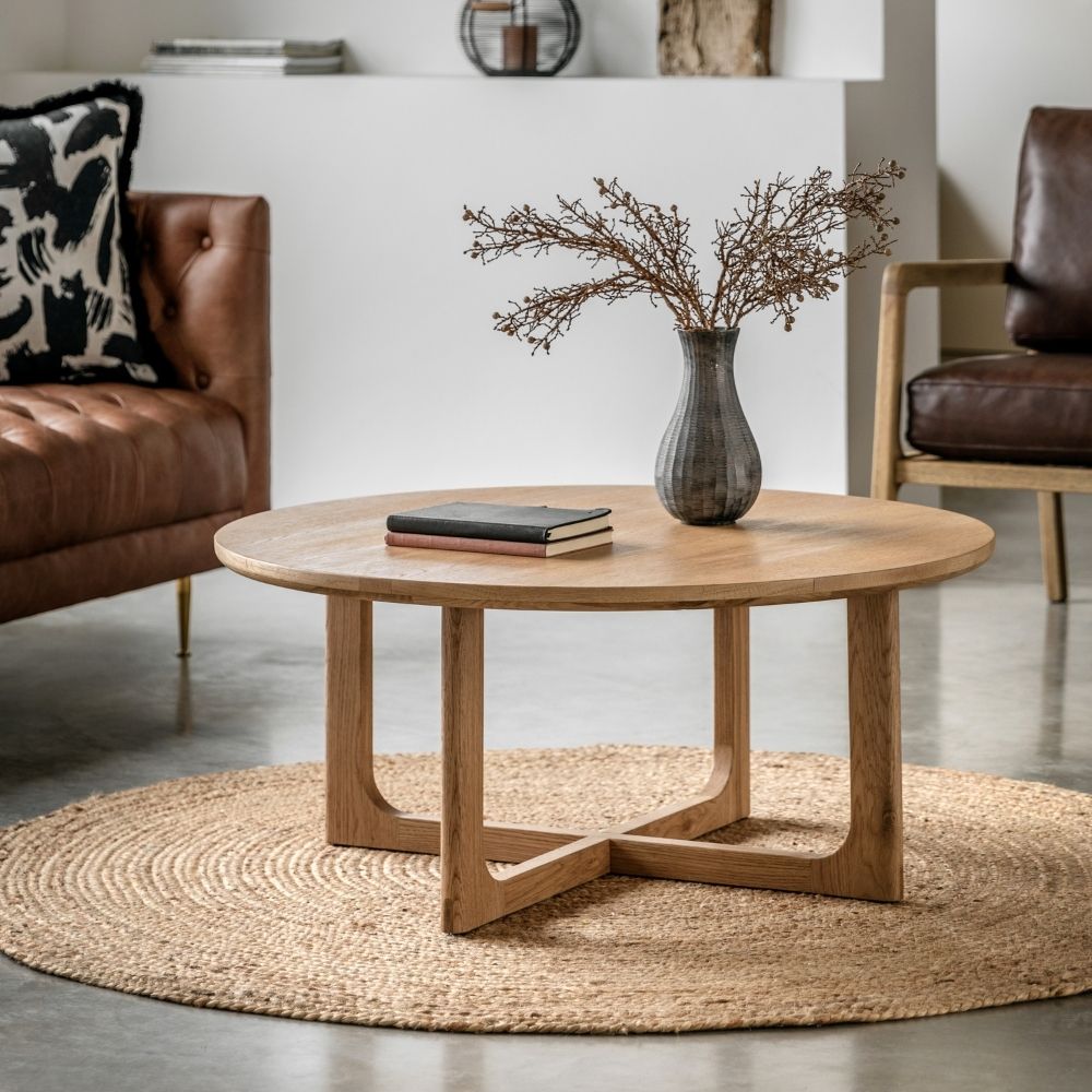 Natural oak round coffee table styled in a modern living room with leather seating, decorative vase and woven rug.