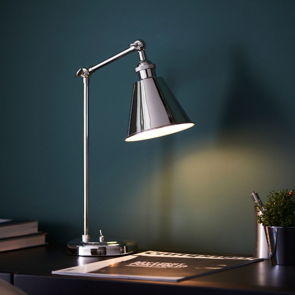 Polished nickel adjustable task table lamp illuminating a modern desk with books and accessories against a dark teal wall.