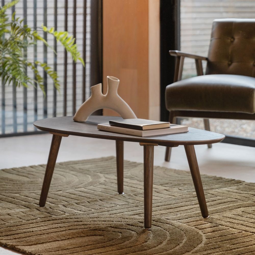 Smoked oak oval coffee table with Scandinavian legs styled in a modern living room with armchair, rug and decorative accessories.