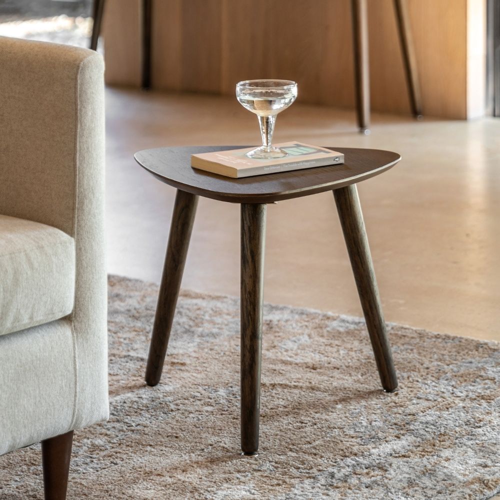 Smoked oak triangular side table with tapered legs styled beside an armchair with a book and glass in a contemporary living room.