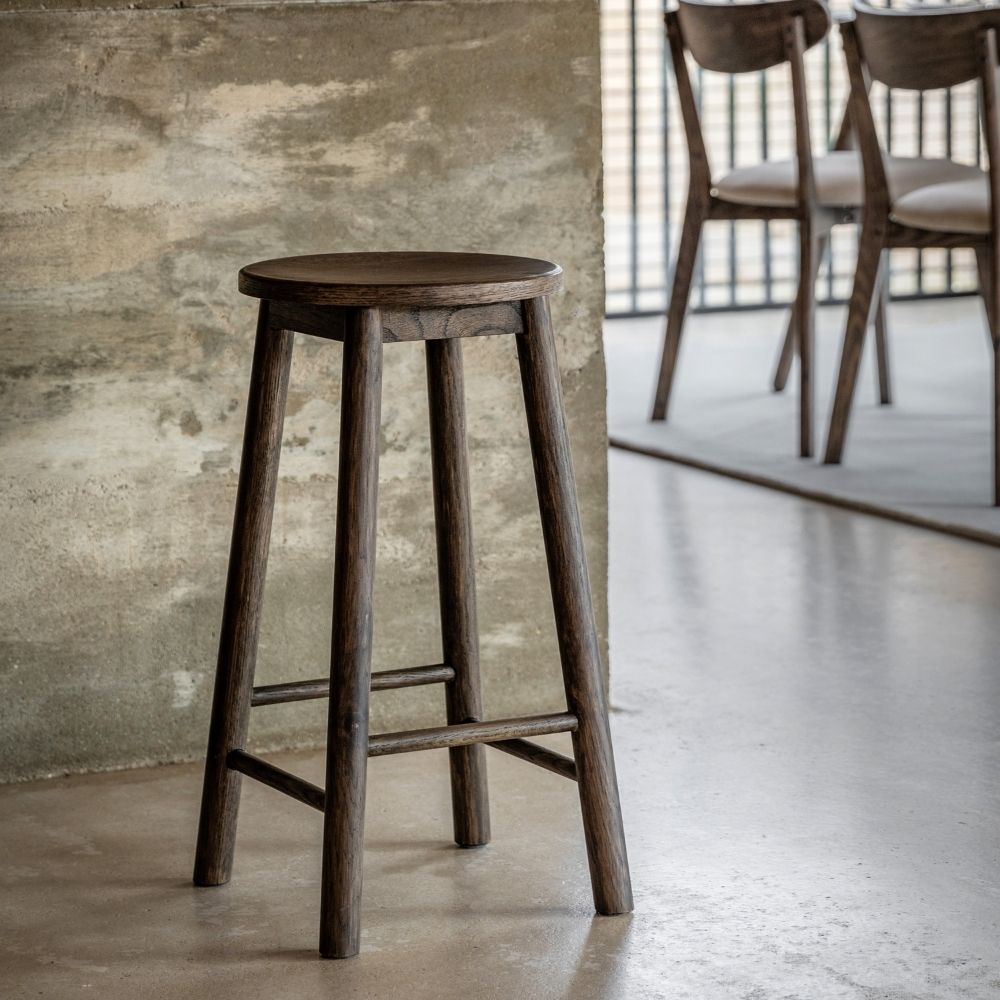 Smoked oak wooden bar stool placed in a modern interior with a concrete wall and dining chairs in the background.