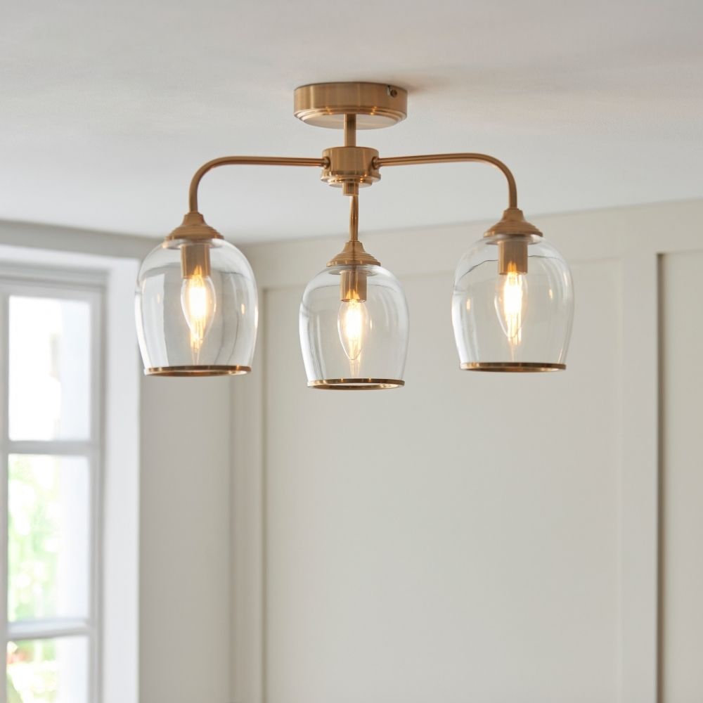 Modern three arm ceiling light in brushed brass with clear glass shades and exposed filament bulbs in a bright interior setting.