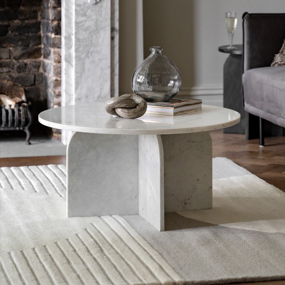 Round white marble coffee table with sculptural base styled in a modern living room with fireplace, armchair and decorative accessories.