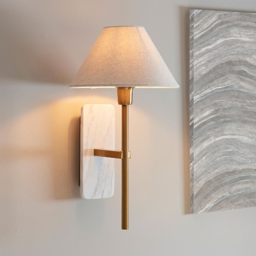 Wall light featuring a white marble backplate with brass arm and neutral linen shade casting a warm ambient glow on a modern interior wall.